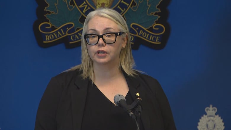 A woman stands at a microphone in front of the RCMP coat of arms.