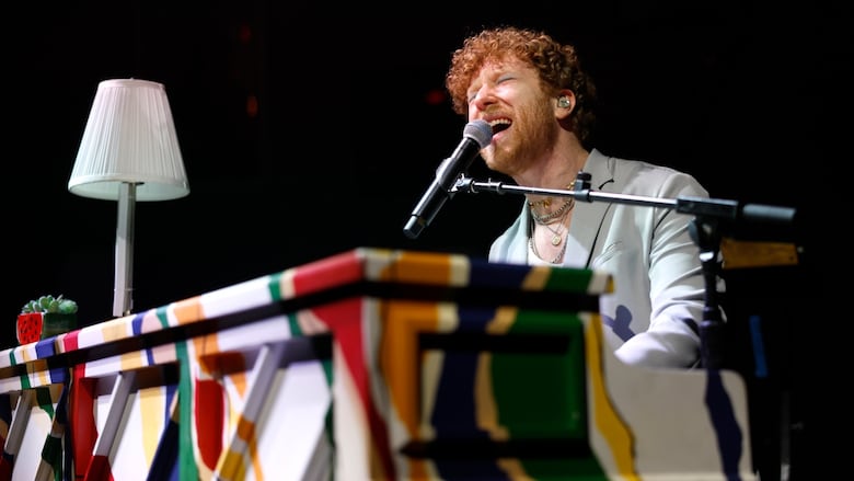 JP Saxe, a white man with curly red hair, sits at a piano and sings.