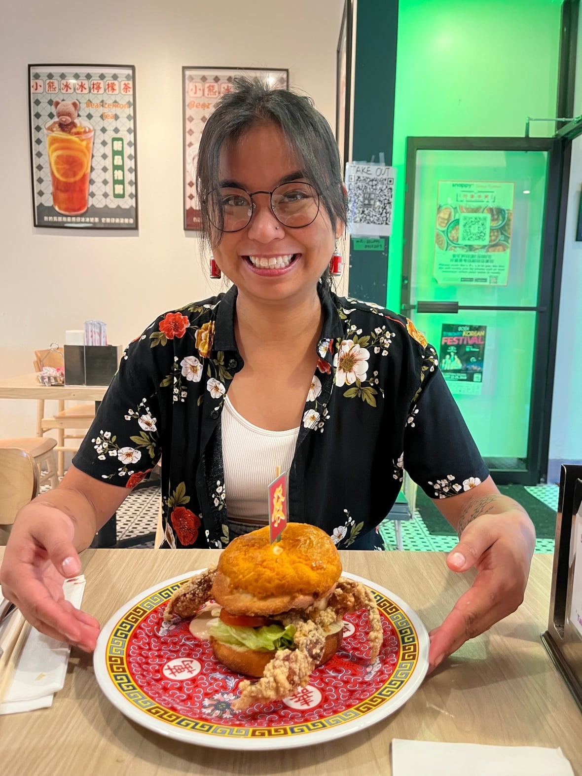 A smiling woman wearing glasses and a black shirt with flowers sits at a restaurant table. She is pointing at a fried sandwich on a red plate in front of her.
