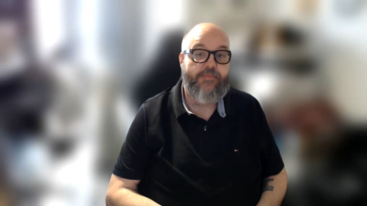 A bald man with a greying beard wearing a black t-shirt and glasses sitting in a chair