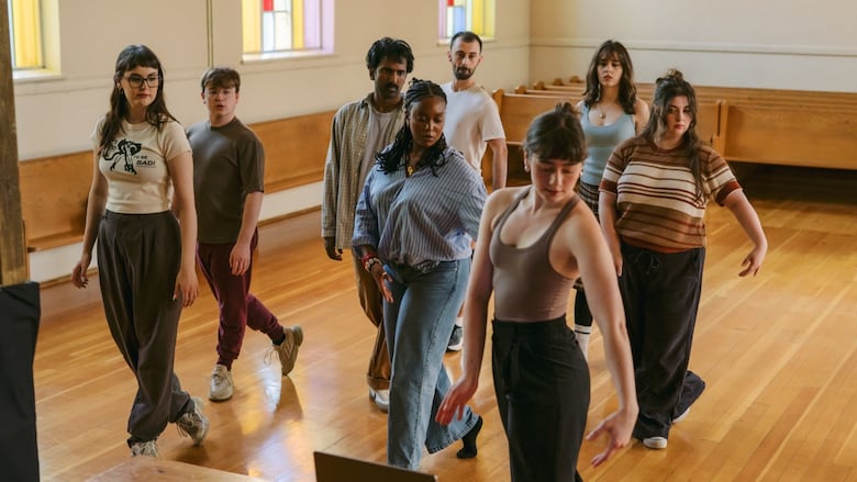 A group of people rehearse a movement in a room with wooden floors, wooden benches and stained glass windows.