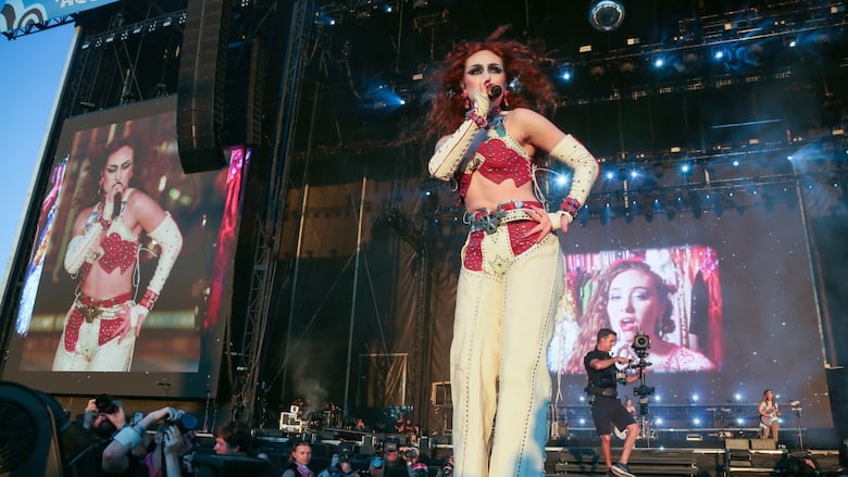 A woman with bright red hair in front of concert stage screens.