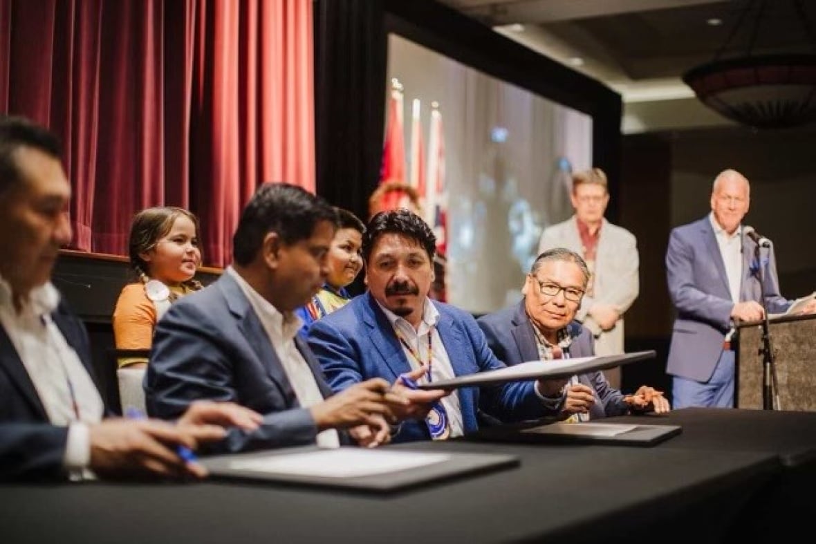 Men sit at a table, passing documents between each others hands, with young people standing behind them. 