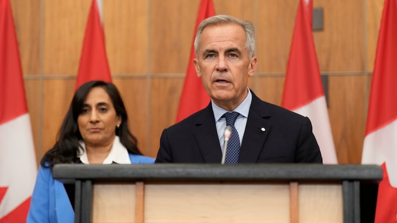 A man in a suit speaks at a podium in front of a row of Canadian flags. A women stands behind him.