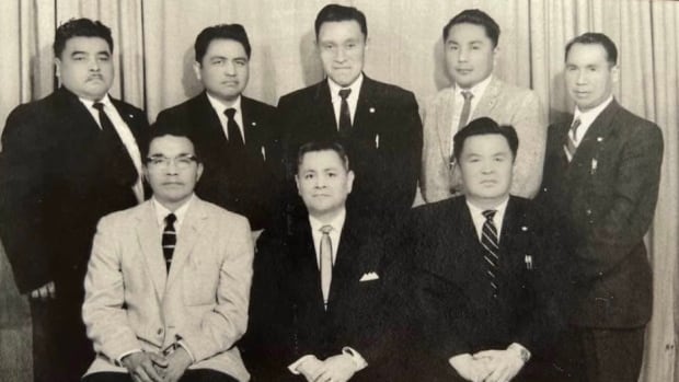 Eight Nisga'a men are wearing suits, lined up in two rows posing for a photo. 