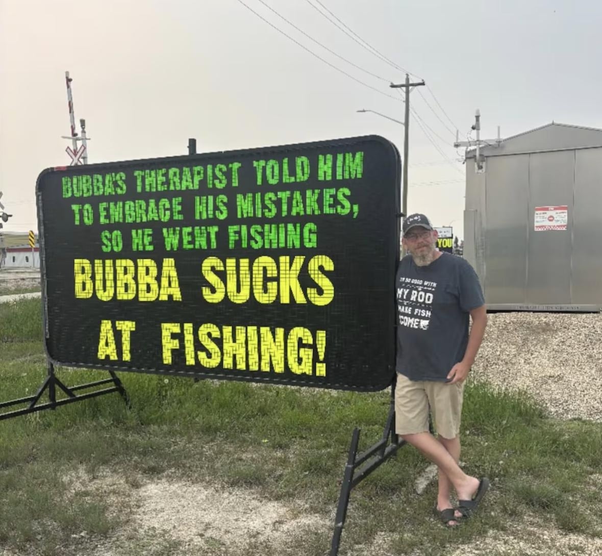 A man leans next to a big sign that reads: Bubba's therapist told him to embrace his mistakes, so he went fishing. Bubba sucks at fishing!