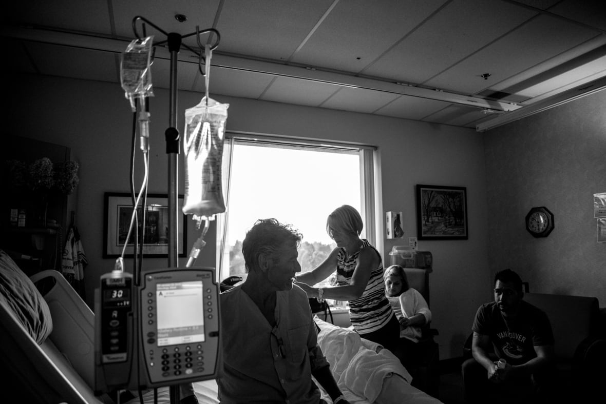 A black and white photo of a man in pyjamas sitting on a hospital bed and surrounded by medical equipment. Three other people are in the room with him.