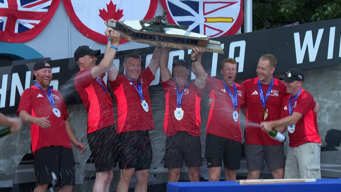 A team of men wearing red uniforms celebrate with a large trophy. They're being sprayed with champagne.