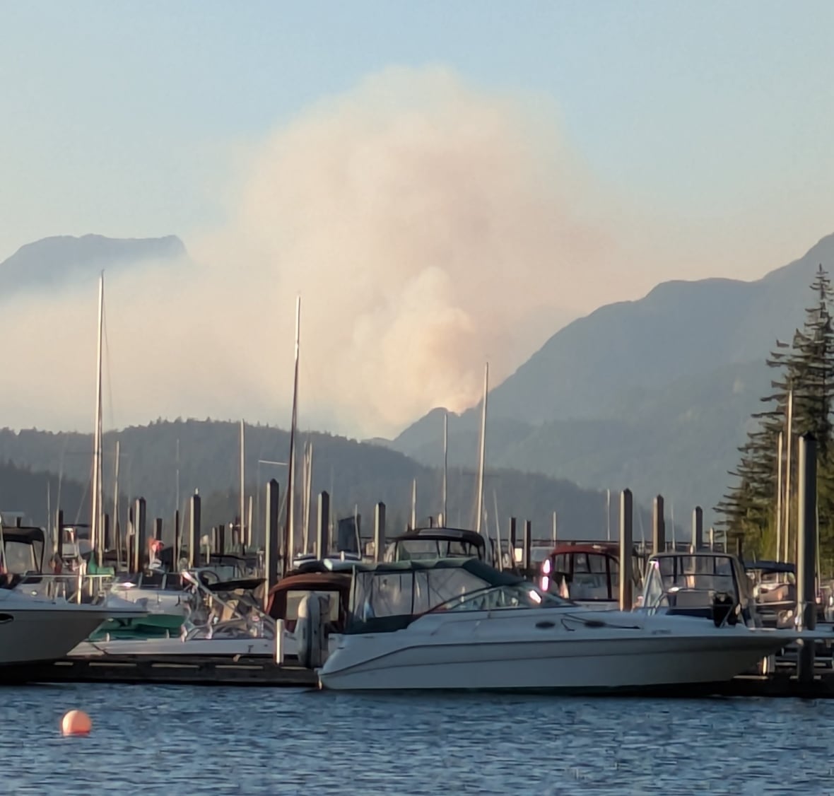 Smoke is seen behind some hills and a row of boats on the water.