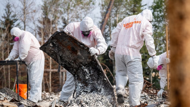 Volunteers wear white suits on rubble as they hold wreckage left from wildfire. 