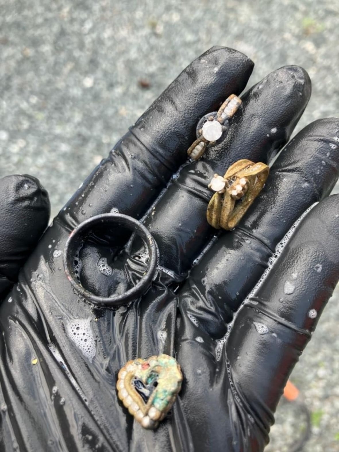 A hand in a black latex glove holding rings and a heart pendant.