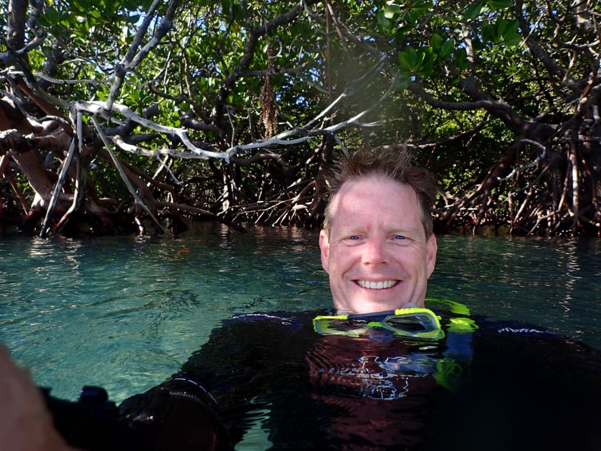 A man in a black wetsuit smiles while submerged in water near some trees.
