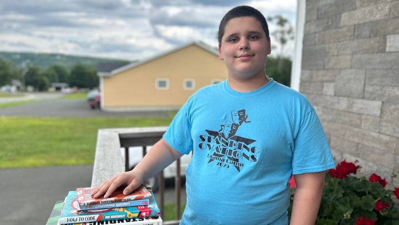 A middle-school aged boy with short black hair smiles at the camera while standing on a front porch with his hand on a stack of books.