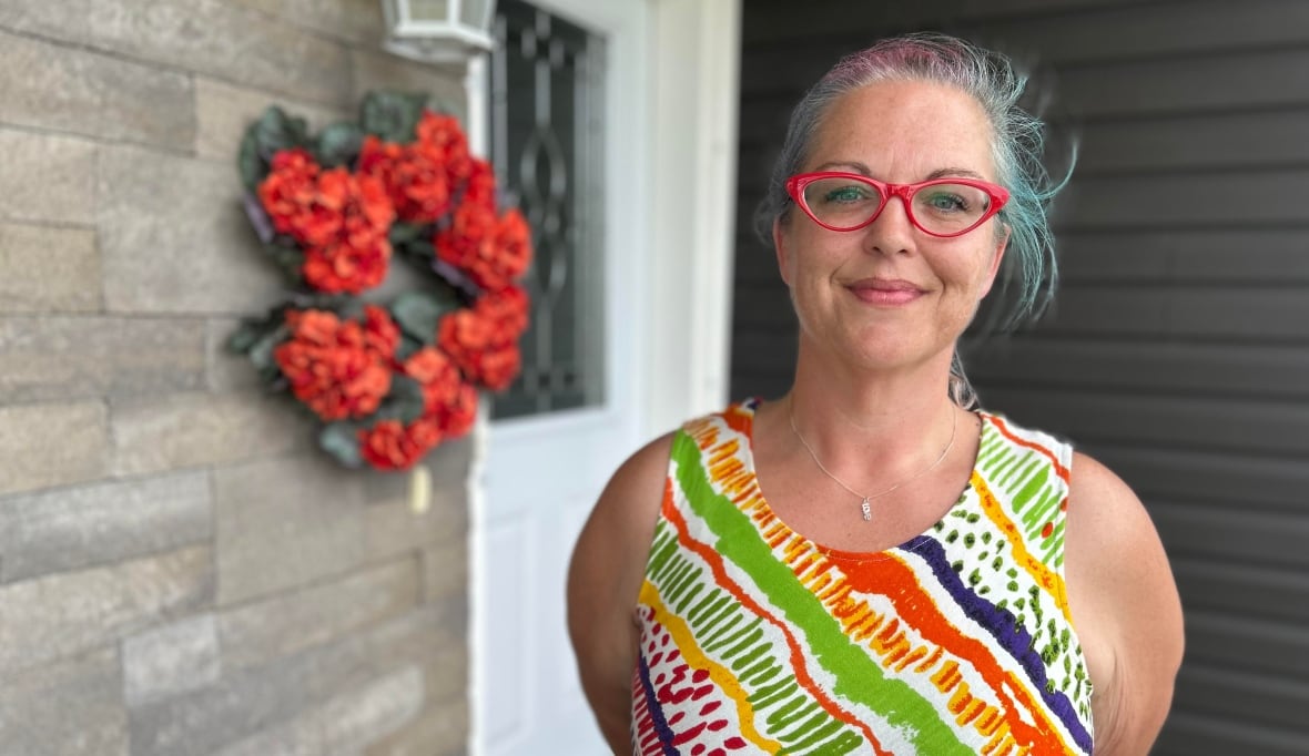 A woman with blue and purple hair pulled back, red glasses, and a colourful sundress, smiles at the camera while standing on a front porch.