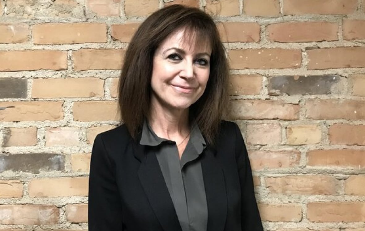 A woman with long dark hair stands against a brick wall. She wears a black blazer over a grey collared shirt.