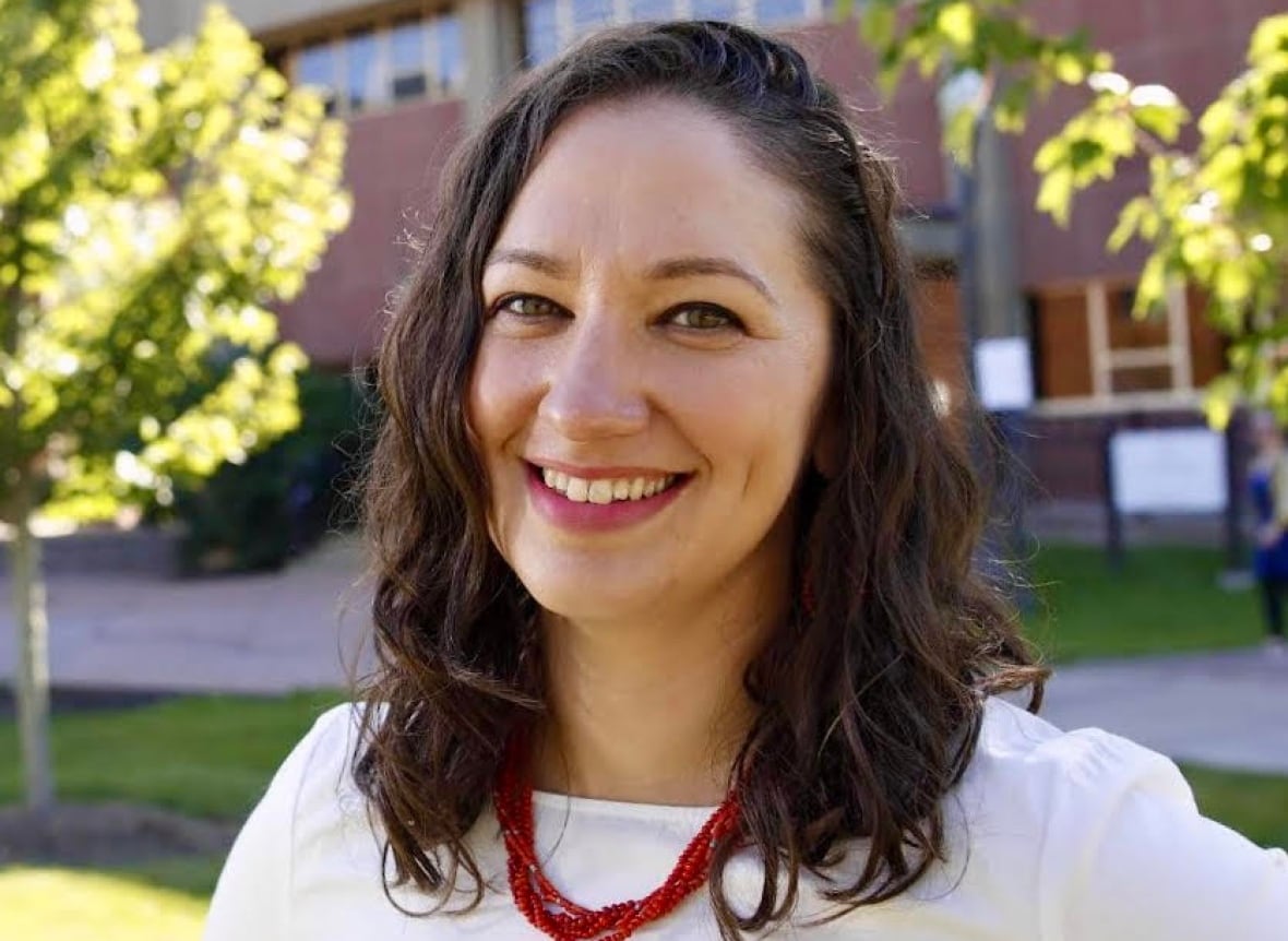 A woman with shoulder-length brown hair, a white shirt and a red, beaded necklace, smiles at the camera with a red brick building in the background.