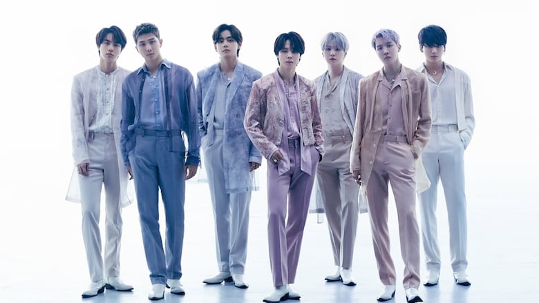 7 men in pastel-coloured outfits stand in a line against a white backdrop, with long shadows extending in front of them.