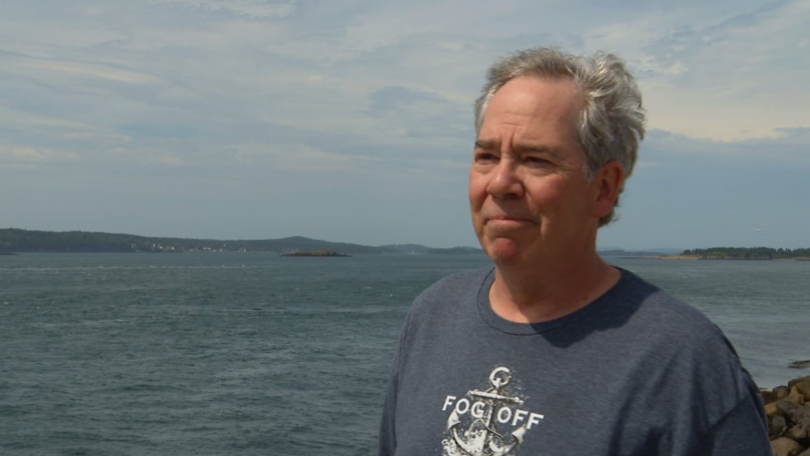 A grinning man wearing a blue t-shirt that says "Fog Off." A bay of water is behind him.