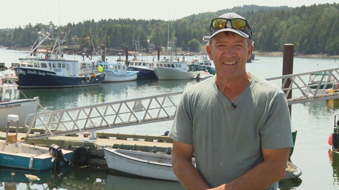 A smiling man wearing a grey t-shirt, a grey hat and sunglasses on the hat, standing in front of a marina.