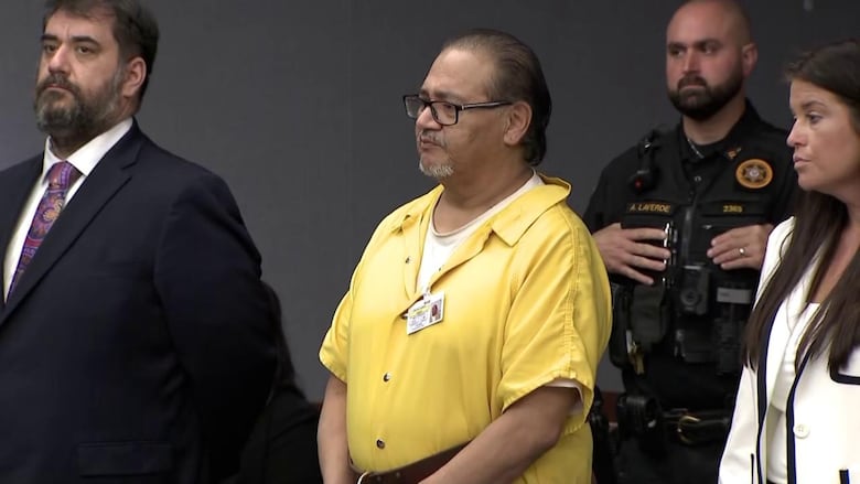 A man in a prison jumpsuit stands, with his lawyers and a police officer near him.