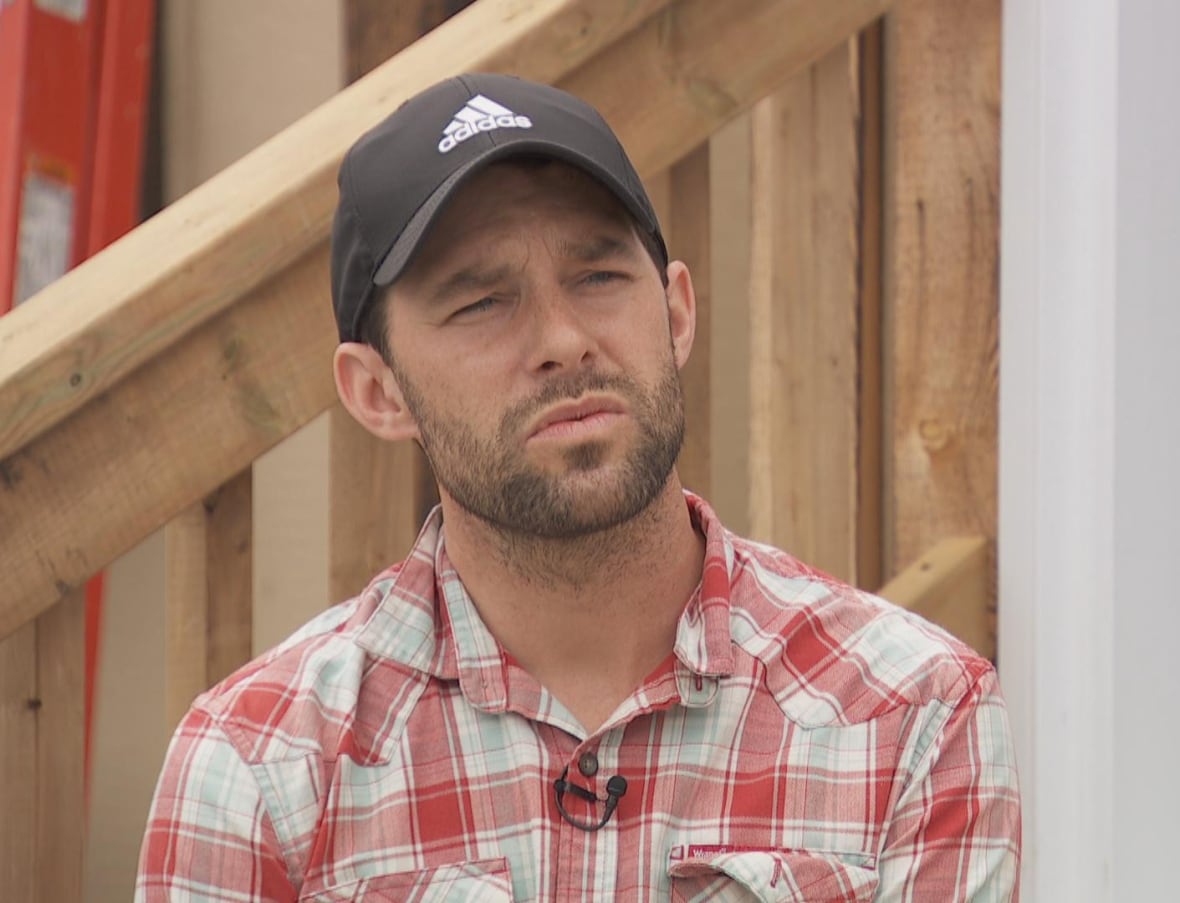 Man, wearing checkered shirt and hat, looking at camera with a neutral expression.