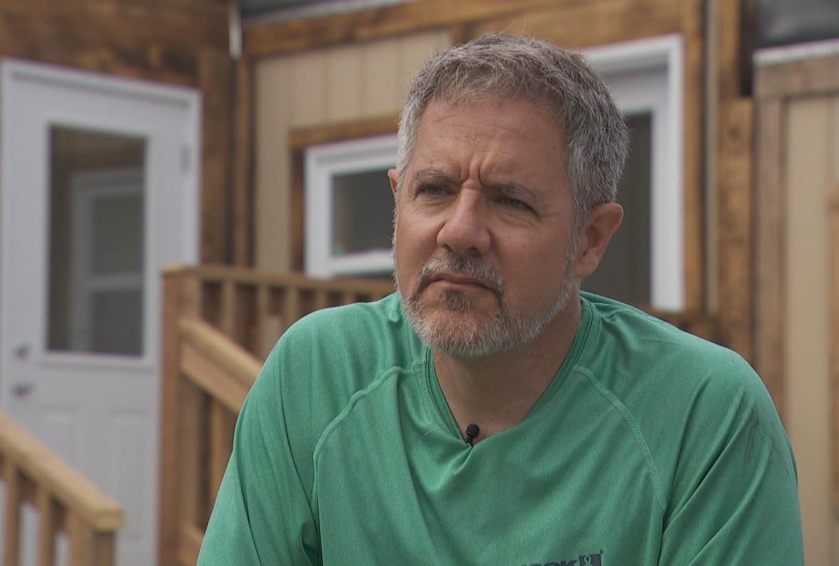 Man with grey hair wearing green shirt sitting on bench looking at camera.