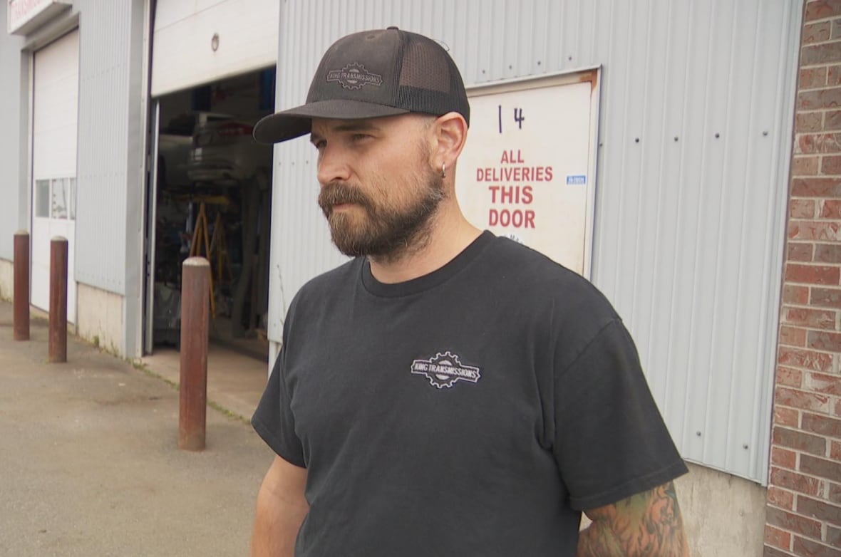 Man, wearing hat, looking at camera on a sunny day with a neutral expression.
