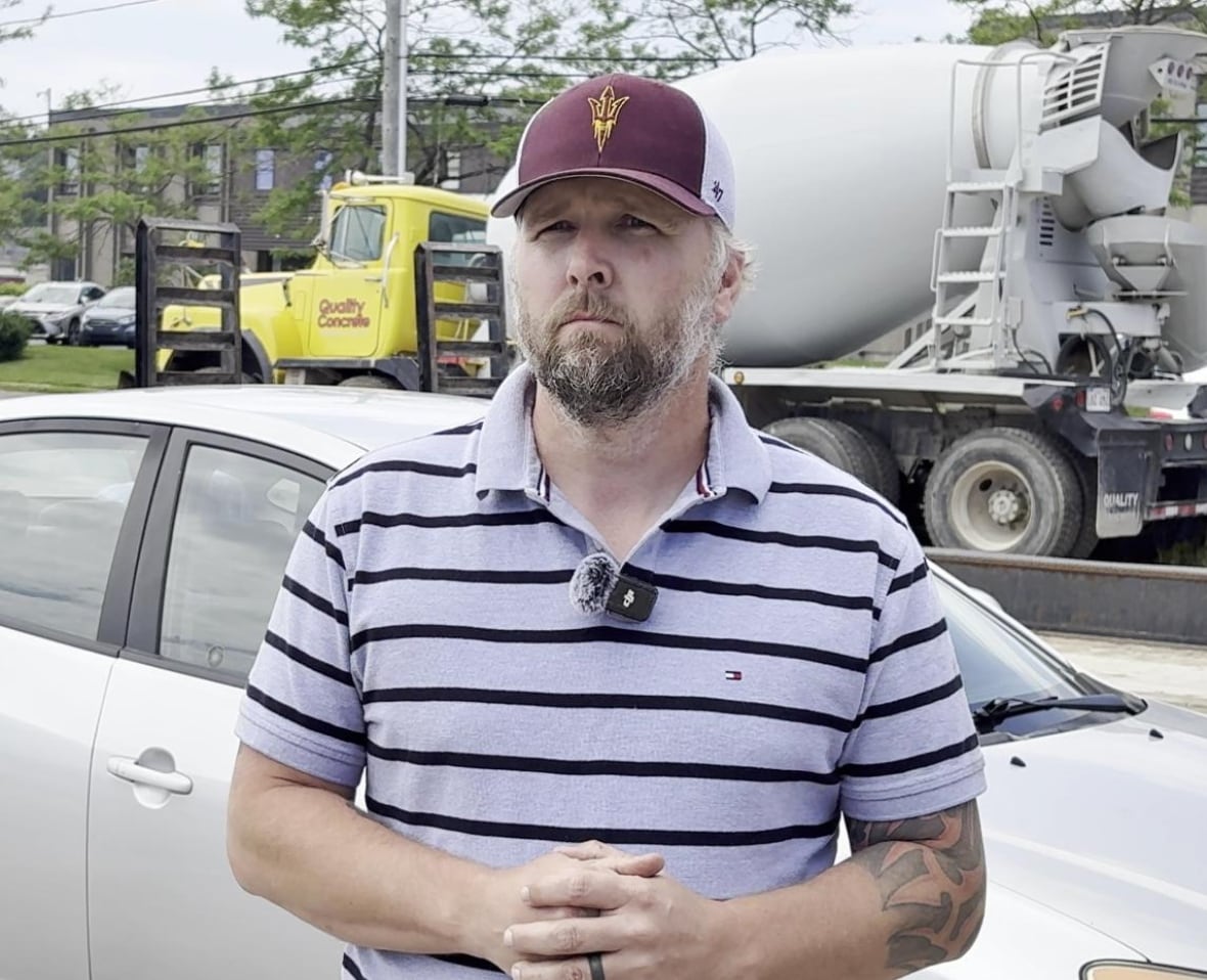 Man, wearing striped t-shirt and hat, looking at camera with a neutral expression with cars passing by behind him.