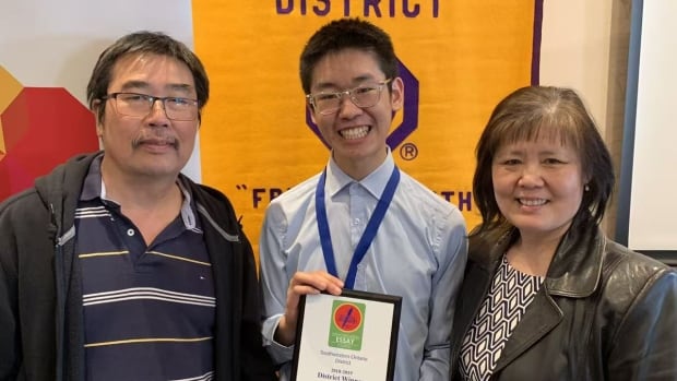 A young man shows off a certificate, flanked by two older people in front of a large poster.