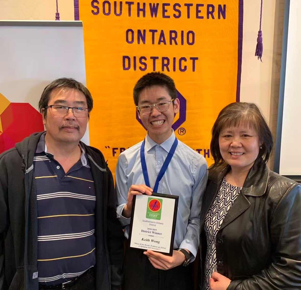 A young man shows off a certificate, flanked by two older people in front of a large poster.