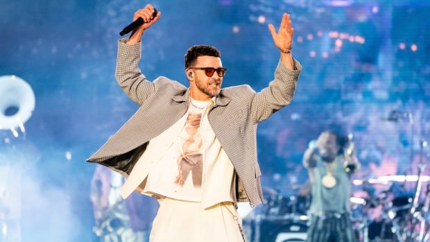 A man wearing sunglass, a grey sports jacket, white shirt and white pants performing on stage, raising his arms in the air with a microphone in his right hand, while a band plays in the background behind him.
