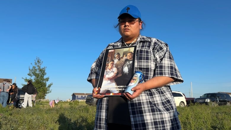 A man looks at the camera while holding a large photo of three men, which says "In loving memory of Trayton Running Bird". He's also carrying a candle with someone's face on it.