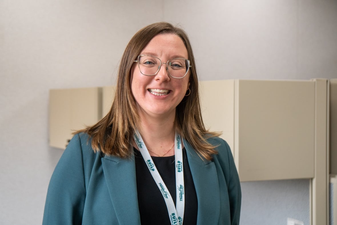 A person wearing a dark teal blazer and black shirt is seen standing in a room, smiling.