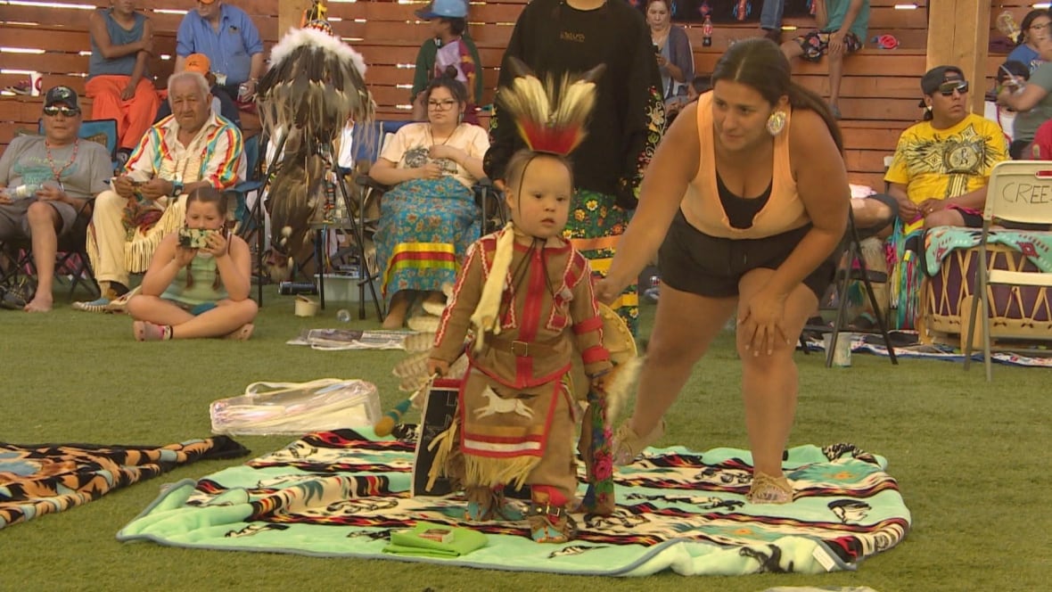 A young boy performs in powwow gear while a woman leans over beside him.