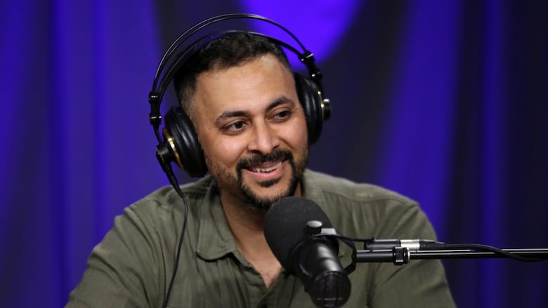 A smiling man wearing over-ear headphones sits in front of a studio microphone.