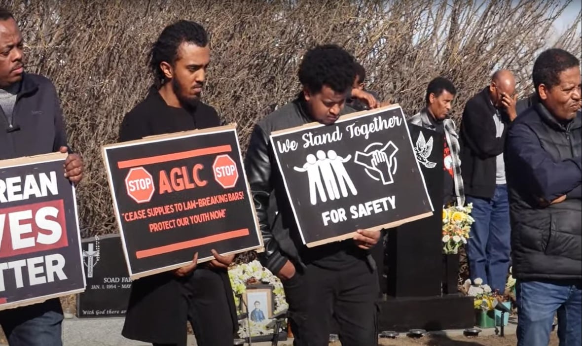 Men hold signs and have grim faces. The signs read Eritrean Lives Matter, AGLC cease supplies to law-breaking bars, and we stand together for safety. 
