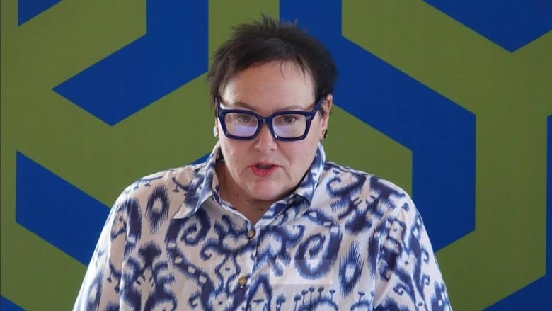 A woman in a blue and white collared shirt with blue framed glasses stands infrom of a blue and green wall.