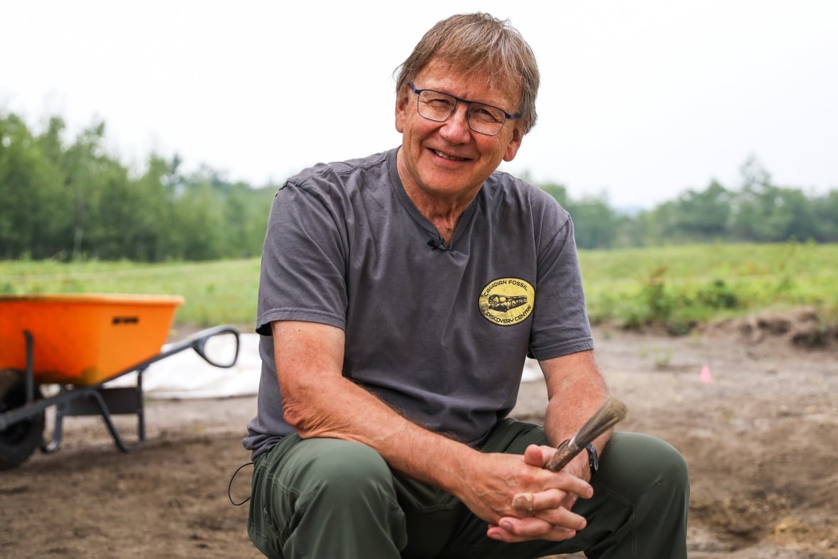 A man in a grey shirt and glasses smiles while sitting next to a fossil dig site.