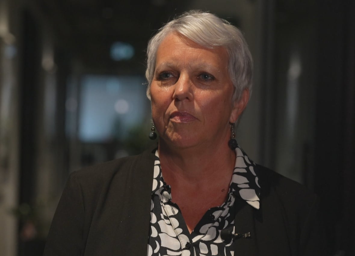 A woman with short silver hair, with a top black and white stamped and a dark blazer, looks out of the left chamber.