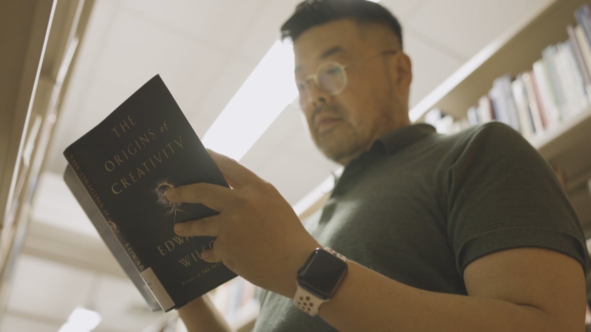 A man with glasses and a short -sleeved pole shirt looks towards an open book in a library.