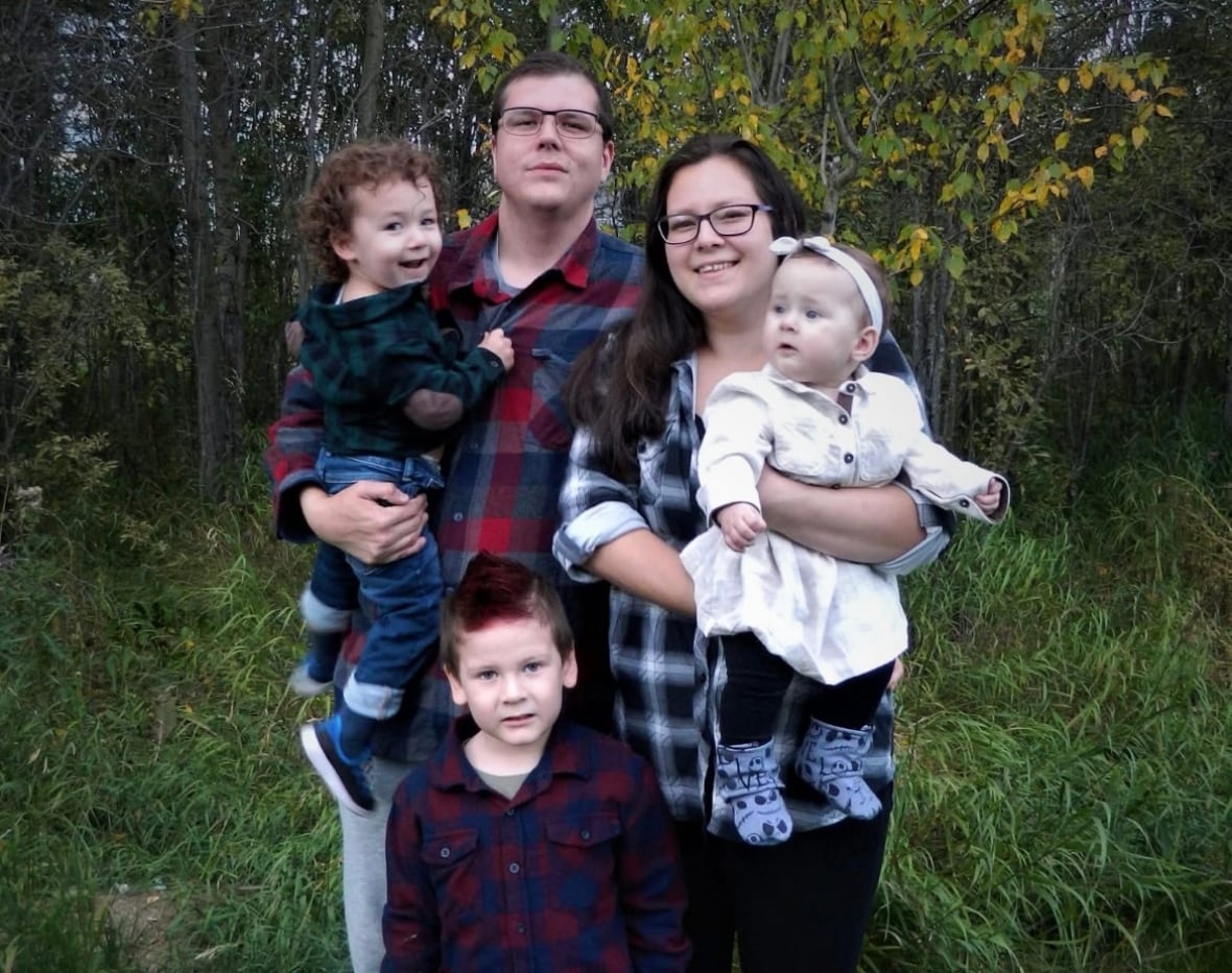 A mother, father and their three children stand in front of a line of trees and other vegetation for a photo.