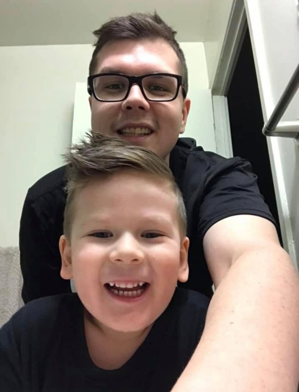 A man in a black T-shirt poses for a selfie with a boy also in a black T-shirt.