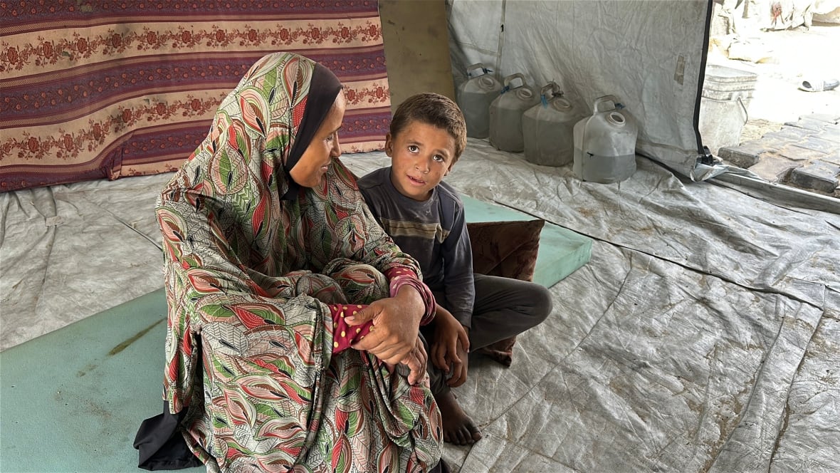 A woman wearing a colourful hijab sits in the middle of a tent looking at a small boy who watches the camera. 
