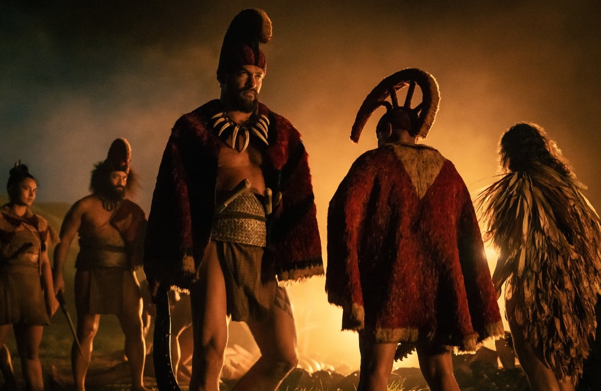 A man wearing a tunic and headress stands in front of a fire.