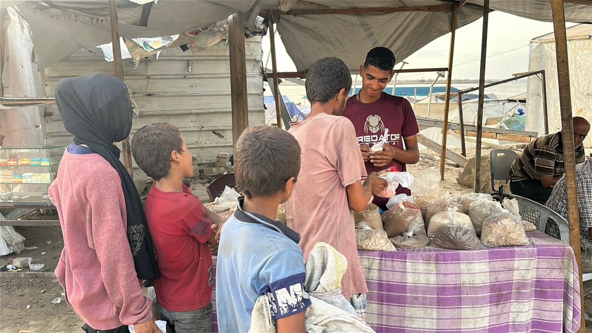 A man and three children stand in front of a makeshift stall where a man counts money behind a table holding small bags of lentils and other foods. 