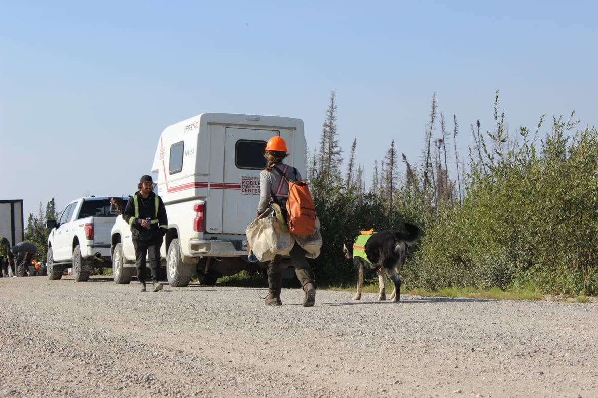 A person and a dog walking along a gravel road to a man and some vehicles.