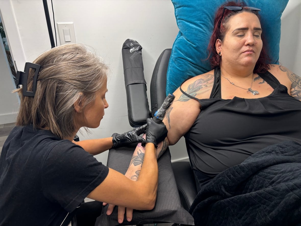 Parnell is tattooing Lenton's right forearm. Parnell is focused on the arm, Lenton has her eyes closed.