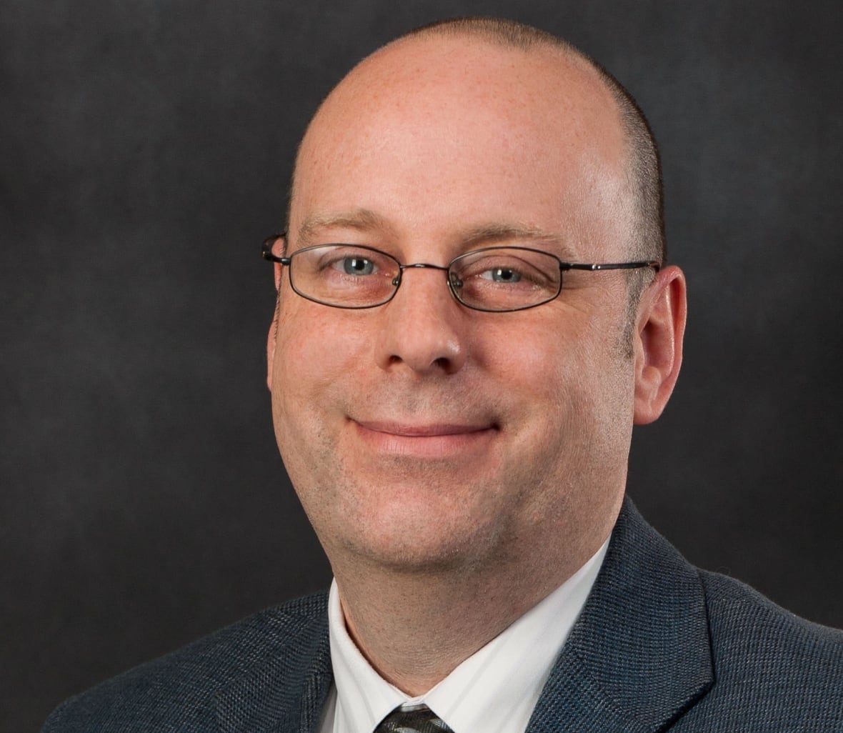 Head-and-shoulders photo of a smiling, balding, bespectacled man in a black jacket, white shirt and tie.