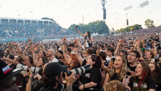 thousands of people are at a concert. you can't see the stage and there's confetti in the air. There are photographers in the foreground. It's daytime.