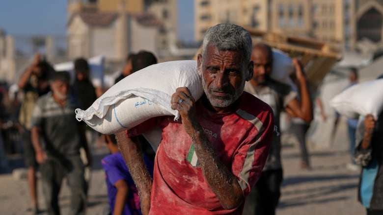 A man carries a white sack over his shoulder. He is covered in dust, and many other people are visible in the background behind them, some also carrying white sacks. A ruined building is visible in the blurred distance.